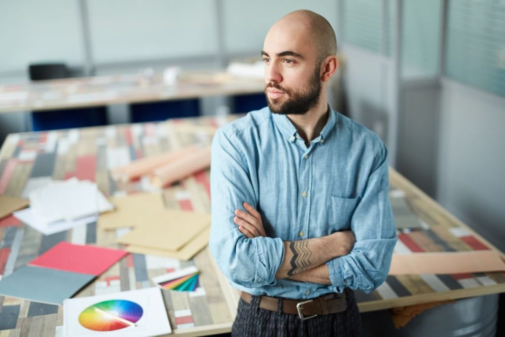 Serious purposeful handsome young bearded man with tattoo leaning on table and looking away while thinking about new design project