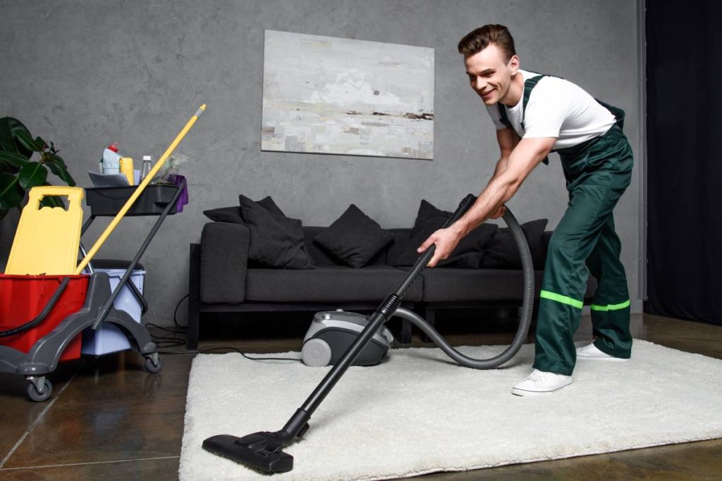 smiling young professional cleaner cleaning carpet with vacuum cleaner