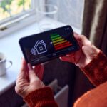 Senior Woman With Smart Meter Trying To Keep Warm By Radiator During Cost Of Living Energy Crisis
